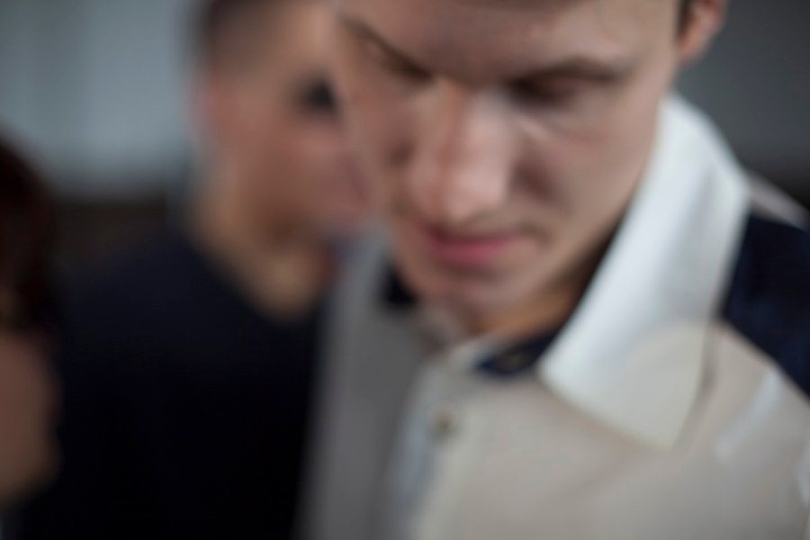 Ein junger Mann steht ganz nah an der Kamera. Sein Kopf ist leicht gesenkt, seine Augen geschlossen. Man sieht sein komplettes Gesicht, allerdings verschwommen, seine Lippen, seine Nase, seine Augenbrauen. Er trägt ein weißes Hemd mit einer schwarzen Schulterklappe und schwarzem Innenkragen. Ein Mann im schwarzen T-Shirt geht hinter ihm vorbei. Der junge Mann hält inne, vielleicht ist ihm etwas eingefallen und er will es nicht vergessen. Oder umgekehrt? Versucht er sich an etwas zu erinnern, was gar nicht mit dieser Situation hier zu tun hat? (Aljoscha Khairetdinov)