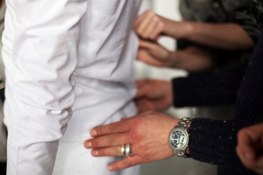 Man sieht einen Mann in Weiß von hinten und vier Hände, die den Stoff ertasten. Zwei Hände im Hintergrund gehören einer Frau. Die zwei Hände im Vordergrund einem Mann. Dieser trägt eine große Uhr und zwei massive silberne Ringe.