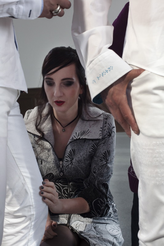 Angeschnittene Silhouetten von zwei weiß angezogenen Männern umrahmen links und rechts dieses senkrechte Foto, auf dem die Hauptfigur eine schlanke schwarzhaarige Frau in einem grau-schwarz gemusterten Kleid ist. Ihr Kleid ist sehr originell geschnitten, mit durchgehendem Reißverschluss und breitem Kragen. Sie kniet vor dem Mann auf der linken Seite und berührt mit der linken Hand sein linkes Knie. Ihre Fingernägel sind dunkel lackiert, der Mund rot geschminkt. Die Männer haben weiße Hosen und weiße Hemden an, die sie locker über die Taille tragen. Die Hemdmanschetten sind mit kleinen türkisfarbenen Punkten bestickt und vielleicht auch schwarz gefüttert. (Ewa Maria Slaska)