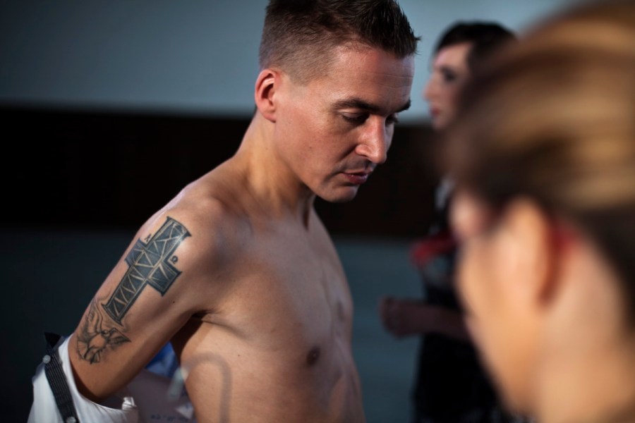 Ein schlanker junger Mann mit kurzen Haaren und entblößtem, unbehaartem Oberkörper. Er ist gerade dabei, sich das Hemd auszuziehen. Er steht im Profil, so dass uns sein rechter Oberarm zugewandt ist, auf dem sich eine markante dunkelblaue Tätowierung befindet. Sie besteht aus einem großen Christuskreuz und darunter einem nicht deutlich zu erkennenden Vogel mit ausgebreiteten Flügeln – vielleicht ein Phönix. (Karl Ahl)