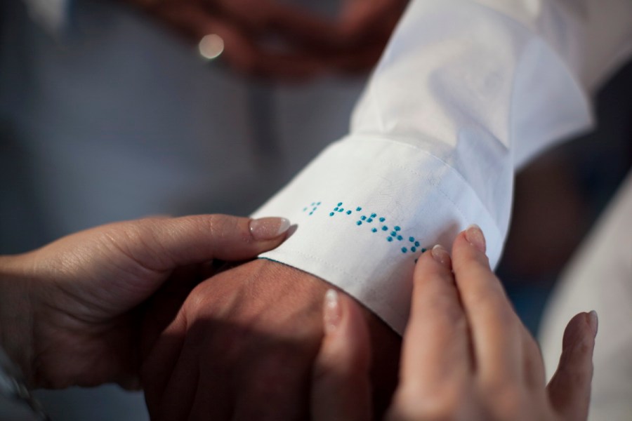 Zwei Frauenhände berühren eine Männerhand. Die Finger der Frau sind manikürt, spitz gefeilt, weiße Ränder, klarlackiert. Ihre eine Hand hält die Hand des Mannes fest, die andere tastet über die Manschette des weißen, gebügelten Hemdes und liest die in türkis gestickte Punktschrift. (Niki Matita)