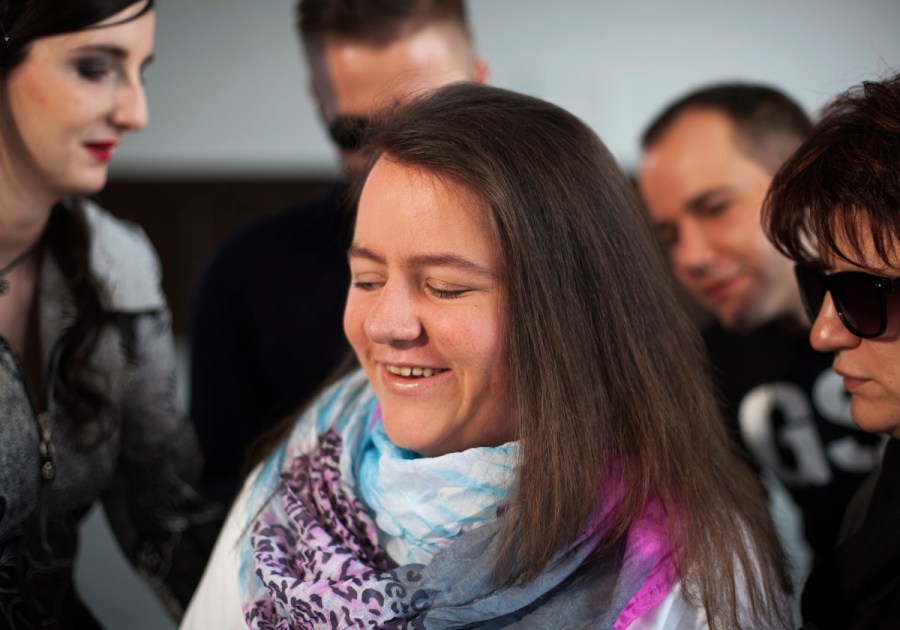 Eine fröhliche Szene. Im Mittelpunkt des Bildes ist eine junge dunkelblonde Frau mit blauem Schal zu sehen. Die Frau schließt die Augen und lächelt sehr vergnügt, eigentlich strahlt sie. Rechts von ihr sieht man eine Frau, schwarz angezogen, mit schwarzer Sonnenbrille. Sie wirkt ein bisschen streng, aber auf keinen Fall unzufrieden. Links von der Frau mit Schal steht eine junge schwarzhaarige Frau in grauem Kleid. Ihr Gesicht ist nur zum Teil sichtbar, man sieht jedoch, dass auch sie lächelt. Im Hintergrund zwei Männer. Einer ist kaum sichtbar, der andere lächelt zufrieden.