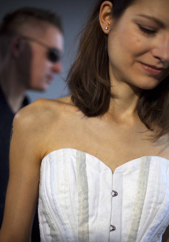 Im Vordergrund, sehr nah, als ob sie direkt vor uns steht, ist eine schöne schlanke Frau. Im Hintergrund, über ihre nackte Schulter hinweg, ist unscharf ein Mann mit Sonnenbrille zu sehen. Beide schauen rechts zur Seite. Die Frau trägt eine weiße Korsage mit grauen Streifen, die in der Mitte markante silberne Verschlüsse hat. Ihr glattes braunes Haar ist nach hinten gestrichen, so dass es ihr linkes Ohr mit einem glitzernden Stein im Ohrläppchen freigibt. Sie neigt ihren Kopf leicht nach unten. Ihre Augen sind geschlossen. (Karl Ahl)