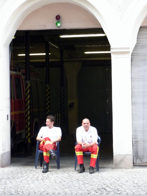 Zwei Feuerwehleute sitzen vor einer Toreinfahrt in der Sonne