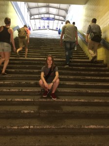 Junge Frau auf Bahnhofstreppe sitzend mit laufenden Menschen im Hintergrund.