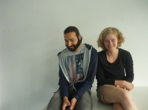 Eine junge Frau und ein junger Mann sitzen auf einem weißen Tisch vor einer weißen Wand.