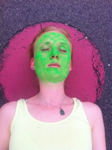 Das grün geschminkte Gesicht einer Frau vor einem rötlichen Hintergrund