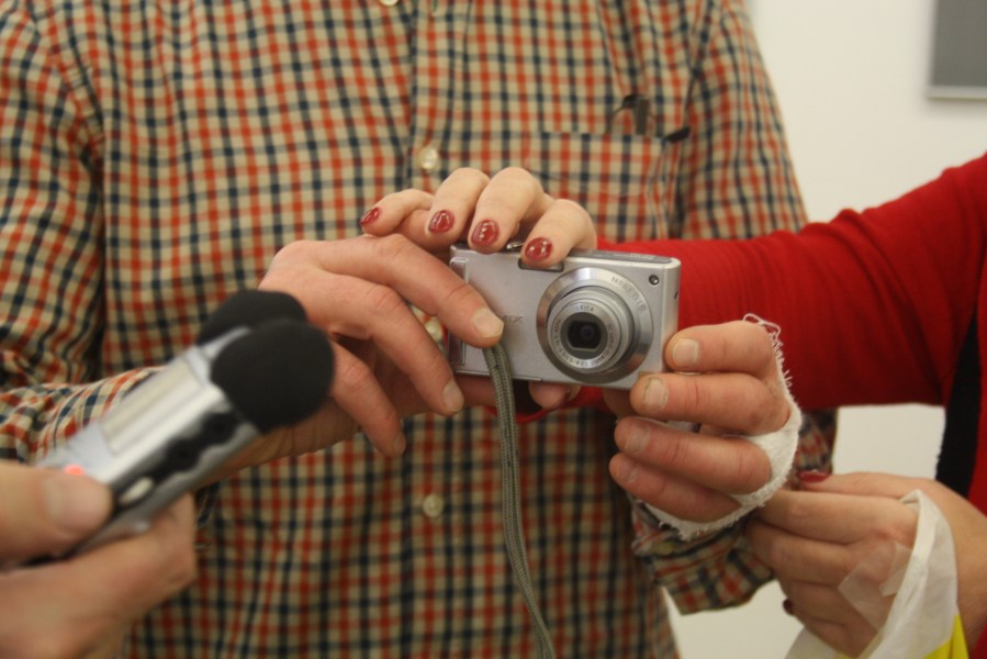 Ein kleiner Fotoapparat in den Händen eines Mannes, die eine Hand ist bandagiert. Die Hände einer Frau betasten die Kamera. Von links wird ein Mikrofon ins Bild gehalten