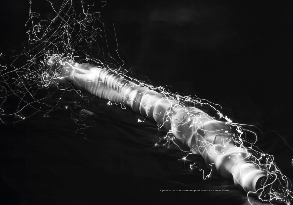 Das Portfolio der Blinden Fotografen in Berlin von 2024/2025.  

Seiten 8 und 9:

Ein doppelseitiges Bild, Silja Korn: “Der Wurm, Lichtübermalung einer Skulptur von Johannes Büttner”

Das querformatige schwarz/weiß-Foto von Silja Korn zeigt die Plastik „ohne Titel“ von Johannes Büttner. Die Plastik ist aus Keramik und hat die Form eines röhren-, bzw. schlauchartigen Objekts. Es ist von einem leicht erhöhten Standpunkt aus aufgenommen, so dass man auf die obere und linke Seite des Objekts sieht. Es erstreckt sich diagonal von links oben nach rechts unten und ist leicht gebogen. Es hat eine unregelmäßige Oberfläche. An einigen Stellen sind ringförmig um den Objektkörper verlaufende Rillen in unterschiedlichen Abständen zueinander, an anderen Stellen sind Einbuchtungen und Dellen.
Das Objekt wird vom schwarzen Untergrund umrahmt. Es liegt auf einem Stoff, der in unregelmäßigen Falten liegt. Der Stoff ist als solcher aber nur in der Nähe des Objekts zu erkennen. Zu den Bildrändern hin verliert er sich im Dunkeln. Bei näherem Hinsehen, sieht man auf der linken Bildhälfte, in der Nähe des Objekts, ein relativ kurzes, durchsichtiges, nur locker zusammengelegtes Kabel liegen sowie einen kleinen quadratischen Apparat und eine kleine durchsichtige Röhre mit einem Schraubverschluss.
Das zentrale Objekt wird von Lichtspuren umspielt. Die Lichtspuren in der linken, oberen Bildhälfte umspielen das Objekt weiträumig und sind vom Bildrand angeschnitten. Auf dem Weg zum unteren, rechten Bildrand hin, läuft die Lichtspur nah an der oberen Seite des Objekts entlang und umspielt es zum unteren rechten Ende hin sehr eng in unregelmäßigen Spuren. Auf den Lichtspuren leuchten einzelne Punkte hell auf. Die Lichtpunkte sind an anderen Stellen zu finden, wie auf dem Farbfoto. Einige der Lichtpunkte sind so hell, dass sie von kleinen Strahlenkränzen umgeben sind. (Bildbeschreibung von Birgit Plinke) 
