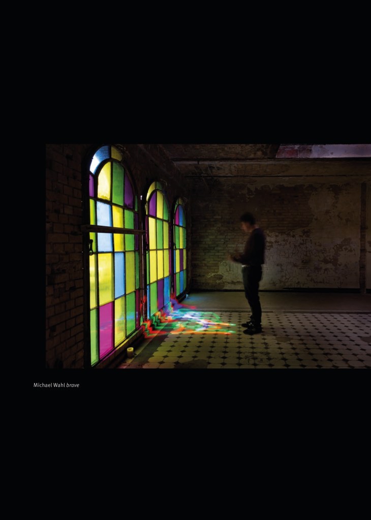 Das Portfolio der Blinden Fotografen in Berlin von 2024/2025.  

Seite 12:

Bild, Michael Wahl:” brave”

Links im Bild drei über mannshohe, oben gewölbte Fenster, die aus Segmenten in verschiedenen Farben zusammengesetzt sind, so dass sie an Kirchenfenster erinnern - violett, grün, blau, gelb. Rechts steht im Profil ein Mensch, zu den Fenstern gewandt, dessen Kontur teilweise mit dem dunklen Hintergrund verschmilzt. Er scheint zu Boden zu blicken. Vor ihm befinden sich sozusagen bunte Lichtpfützen, die nach Form und Farbe wirken, als würde die Farbe aus den Fenstern auf den gefliesten Fußboden fließen. 

