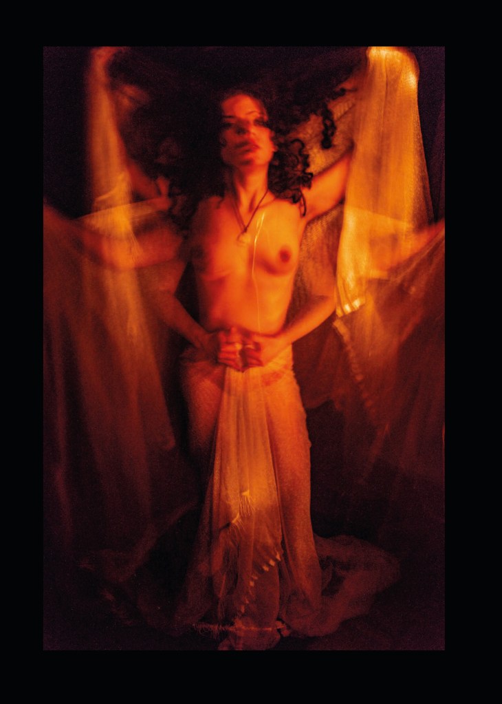 Das Portfolio der Blinden Fotografen in Berlin von 2024/2025.  

Seite 15:

Ein ganzseitiges Bild, Mary Hartwig: “Shivani”

Eine junge Frau mit langem lockigem Haar, das Kinn selbstbewusst erhoben, fest auf dem Boden stehend und unbekleidet bis auf einen Schleier. Diesen hält sie in drei oder vier verschiedenen Positionen, so dass sich der Eindruck ergibt, sie habe sechs bzw acht Arme. Am prägnantesten ist die mittlere Position, in der sie den Schleier fest, mit beiden Fäusten vor ihrem Nabel zusammenhält. Die weiteren Positionen ihrer Arme sind seitlich, auf unterschiedlicher Höhe, aber immer symmetrisch. Die sich an ihren Seiten überlagernden, teilweise durchsichtig wirkenden Phasen des ausgebreiteten Schleiers könnte man aus der Ferne auch für Engelsflügel in Bewegung halten.
